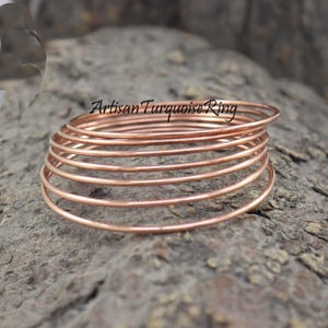 May include: A stack of copper-colored rings, each a thin band of metal. The rings are arranged in a spiral, resting on a textured, gray surface. The text "Artisan Turquoise Ring" is visible in the image.