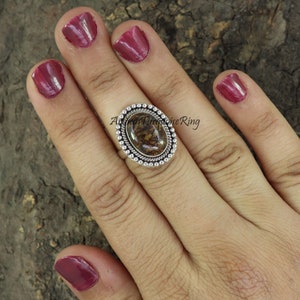 Op de afbeelding: Een zilveren ring met een bruine en witte ovale steen gezet in een geperlde bezel. De ring zit op een vinger met roze nagellak.