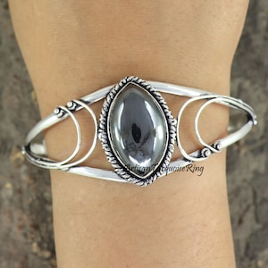 May include: A silver bracelet with a large, oval-shaped, dark gray gemstone set in a rope-like border. The bracelet has a delicate, openwork design with two curved bars.