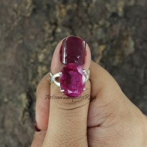 May include: A silver ring featuring a large, oval ruby gemstone. The ring is worn on a finger with dark red nail polish.