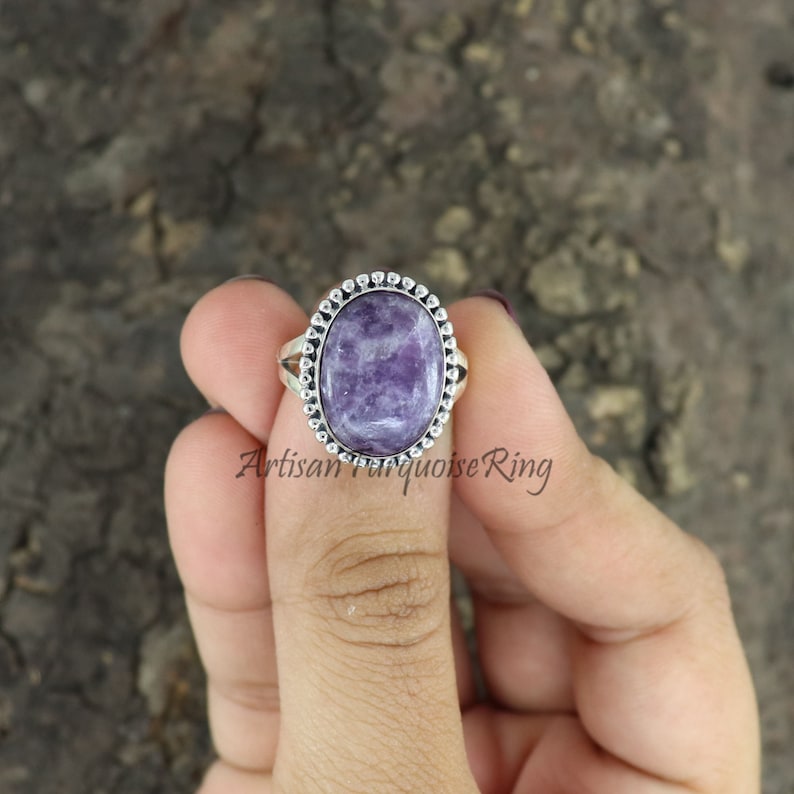 K&ouml;nnte beinhalten: Ein silberner Ring mit einem violetten Edelstein, der in einer perlenbesetzten ovalen Fassung sitzt. Der Ring befindet sich an einem Finger.