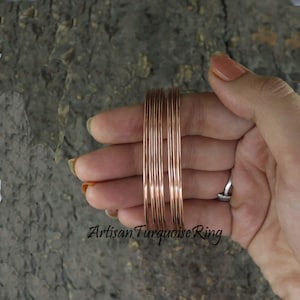 May include: A set of thin, copper-colored bangles. The bangles are stacked together and held in a hand. The text "Artisan Turquoise Ring" is visible at the bottom of the image. The bangles are a simple, elegant design.