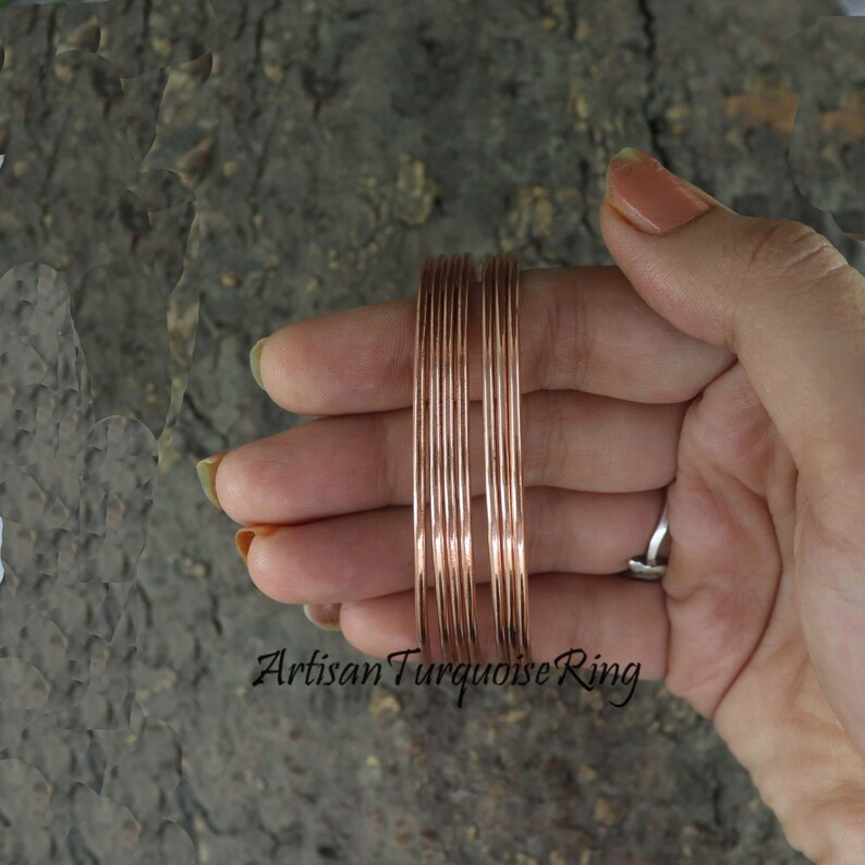 May include: A set of thin, copper-colored bangles. The bangles are stacked together and held in a hand. The text "Artisan Turquoise Ring" is visible at the bottom of the image. The bangles are a simple, elegant design.