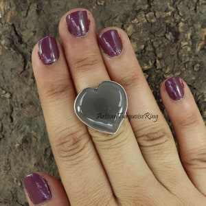 May include: A heart-shaped ring with a gray stone set in a silver band. The ring is on a person's finger.