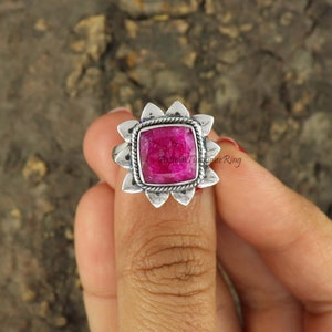 May include: A silver ring with a pink gemstone set in a square shape. The ring has a decorative design around the gemstone.