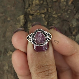 以下が含まれることがあります： ねじれたシルバーバンドにピンクの宝石がセットされたシルバーリング。リングは指にはめられています。