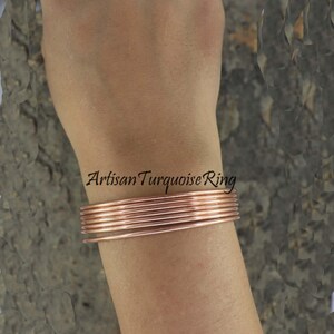 May include: A stack of copper-colored wire bracelets on a wrist. The bracelets are thin and round, with a slight sheen. The text "Artisan Turquoise Ring" is visible on the arm.