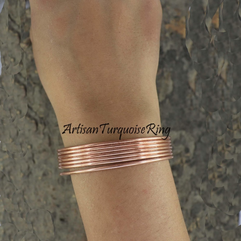 May include: A stack of copper-colored wire bracelets on a wrist. The bracelets are thin and round, with a slight sheen. The text "Artisan Turquoise Ring" is visible on the arm.