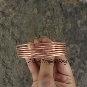 May include: A stack of copper-colored bangles. The thin, round bracelets are stacked together, with the text "Artisan Turquoise Ring" in a script font. The bangles are held against a textured, dark background.
