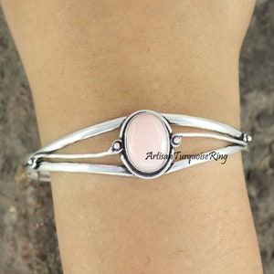 May include: A silver bracelet with a pink oval gemstone set in a silver bezel. The bracelet has three bands and is accented with small silver beads.