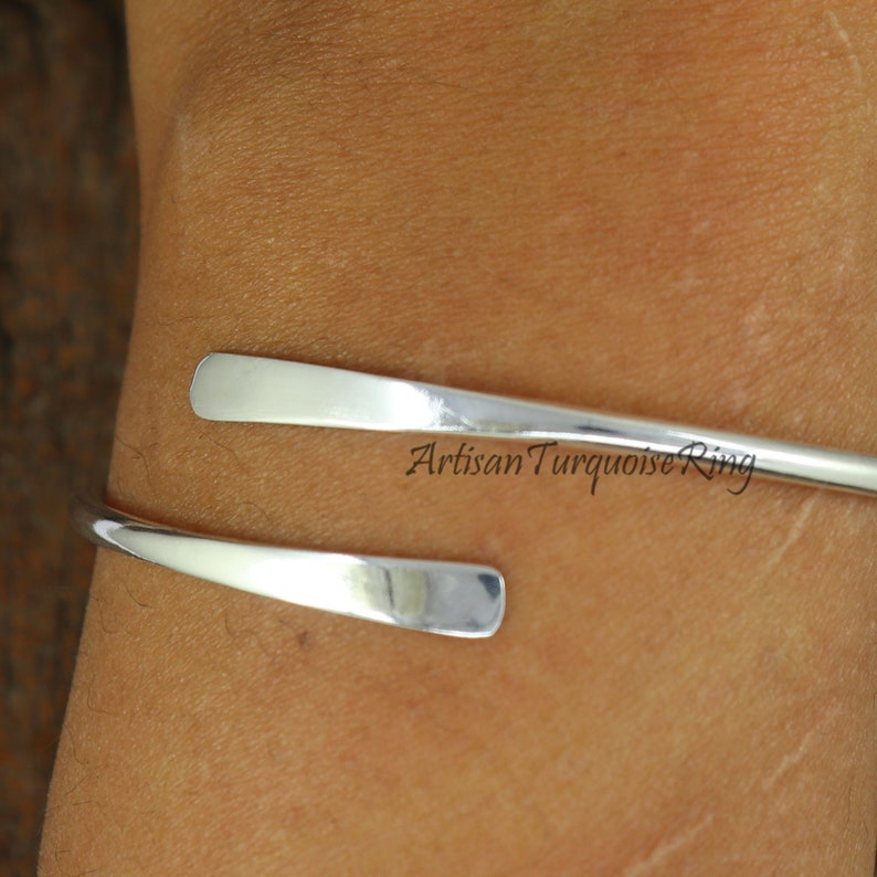 Puede incluir: Un brazalete de plata con un dise&ntilde;o simple y curvo. El brazalete est&aacute; abierto y tiene una banda plana y ancha.