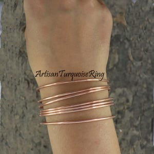 May include: A stack of copper-colored wire bracelets on a wrist. The bracelets are thin and round, with a slight sheen. The text "Artisan Turquoise Ring" is visible in the image.
