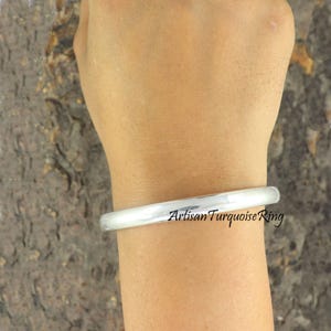 May include: A silver bangle bracelet with the text "Artisan Turquoise Ring" is worn on a wrist. The bracelet has a smooth, rounded design. The background is a textured, neutral colour.