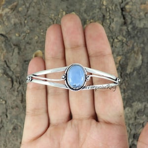Peut inclure: Un bracelet manchette argenté avec une pierre précieuse ovale bleu clair. Le bracelet a un design divisé avec des accents décoratifs et le texte "Artisan Turquoise Ring".
