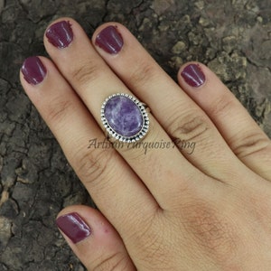 K&ouml;nnte beinhalten: Ein silberner Ring mit einem ovalen, violetten Edelstein, der in einer Perlenfassung sitzt. Der Ring befindet sich an einer Hand mit dunkelviolettem Nagellack.