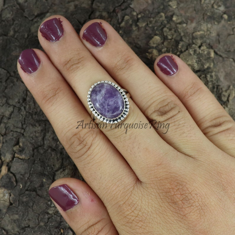 K&ouml;nnte beinhalten: Ein silberner Ring mit einem ovalen, violetten Edelstein, der in einer Perlenfassung sitzt. Der Ring befindet sich an einer Hand mit dunkelviolettem Nagellack.