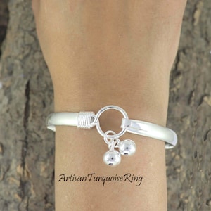 Puede incluir: Un brazalete de plata con cierre de gancho y dos pequeños dijes colgantes en forma de bola. El brazalete se lleva en la muñeca. El texto "Artisan Turquoise Ring" está en la parte inferior de la imagen.