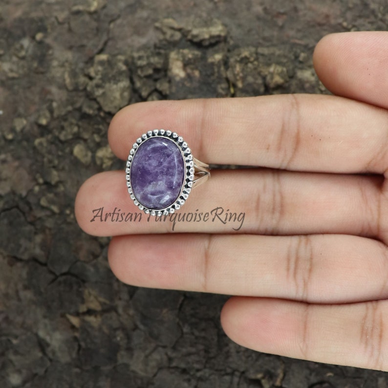 K&ouml;nnte beinhalten: Ein silberner Ring mit einem violetten ovalen Edelstein, der in einer Perlenfassung sitzt. Der Ring befindet sich an einem Finger.