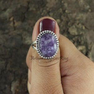 K&ouml;nnte beinhalten: Ein silberner Ring mit einem ovalen, violetten Edelstein, der in einer Perlenfassung sitzt. Der Ring befindet sich an einem Finger.