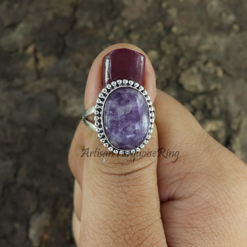 K&ouml;nnte beinhalten: Ein silberner Ring mit einem ovalen, violetten Edelstein, der in einer Perlenfassung sitzt. Der Ring befindet sich an einem Finger.