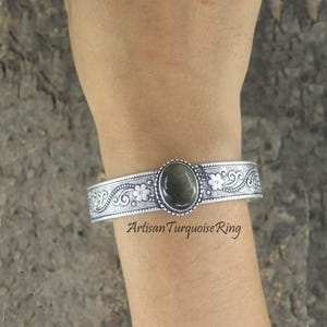 Könnte beinhalten: Silberfarbenes Armreif-Armband mit einem ovalen, olivgrünen Stein in der Mitte. Das Armband hat Blumen- und Rankenmuster. Der Text "Artisan Turquoise Ring" ist sichtbar.