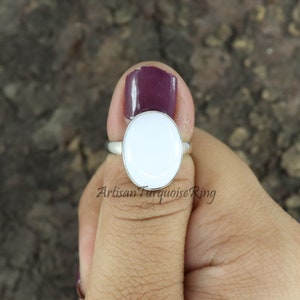 May include: A silver ring featuring a large, oval, white gemstone. The ring is held by a person with dark purple nail polish. The text "Artisan Turquoise Ring" is visible at the bottom of the image.