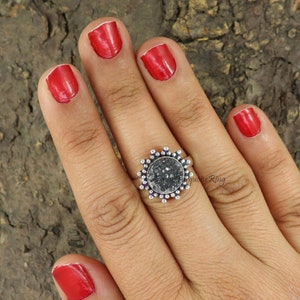 Peut inclure: Une bague en argent avec une pierre ronde noire scintillante sertie dans un motif en forme de soleil. La bague est portée au doigt.