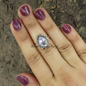 May include: A silver ring with a pear-shaped iridescent gemstone set in a beaded halo. The ring is on a hand with dark red nail polish.