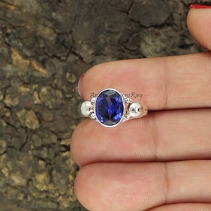 Op de afbeelding: Een zilveren ring met een grote ovale blauwe edelsteen in het midden. De ring heeft twee kleinere zilveren ballen aan weerszijden van de edelsteen.