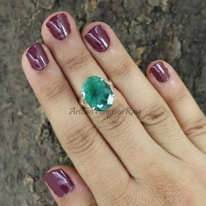 May include: A silver ring with a large, oval-shaped green emerald gemstone. The ring is set with small silver prongs around the stone. The ring is on a person's finger.