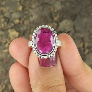 Op de afbeelding: Een zilveren ring met een grote, ovale, gefacetteerde robijn-edelsteen. De ring heeft een geperlde rand rond de edelsteen.
