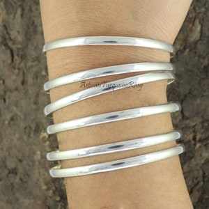 May include: Five silver bangle bracelets stacked on a person's wrist. The bracelets are smooth and shiny.