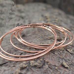 May include: Several thin, copper-colored rings are arranged on a textured, gray surface. The rings are circular and appear to be made of metal. The text "Artisan Turquoise Ring" is visible in the upper left corner.