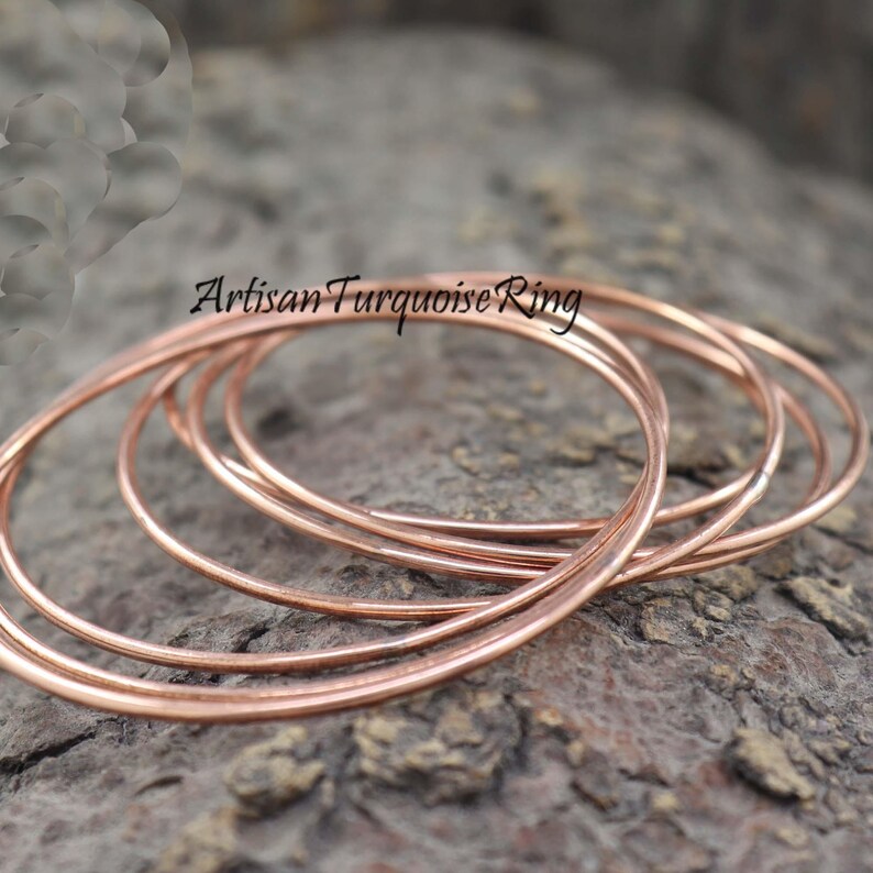 May include: Several thin, copper-colored rings are arranged on a textured, gray surface. The rings are circular and appear to be made of metal. The text "Artisan Turquoise Ring" is visible in the upper left corner.
