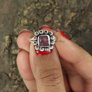 May include: A silver ring featuring a square, reddish-pink gemstone at its centre, encircled by a floral-like pattern of small, circular details. The ring is held by a hand with red nail polish.