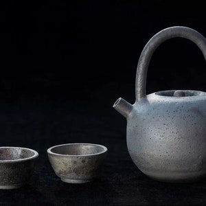 May include: A grey ceramic tea set comprising a rounded teapot with a large handle and spout, and two small cups. The speckled ceramic set is displayed against a black backdrop, creating a minimalist aesthetic.