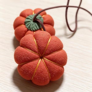 May include: Two fabric pumpkin-shaped ornaments in a vibrant orange hue, featuring segmented sections defined by thin gold thread stitching. The top of each pumpkin has a gathered green fabric stem and a thin brown cord attached for hanging.