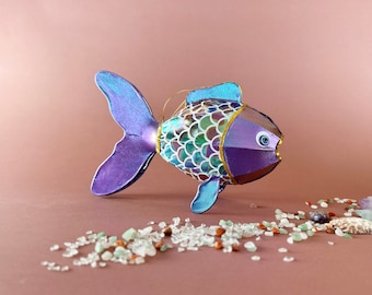 May include: A purple and blue fish ornament with iridescent scales and gold accents. The fish has a white eye with a blue pupil and is sitting on a pink surface with white and brown stones.
