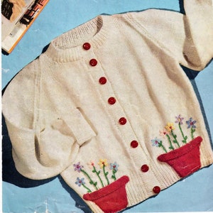 Pode incluir: Um cardigan branco de malha com botões vermelhos e vasos de flores bordados na frente. O padrão é para as idades de 2-3, 3-4, 4-5, 5-6 anos. Marriner's Knitting Wools No. 367.
