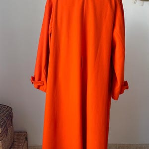 May include: Bright orange long coat with a classic design. The coat features a wide collar, long sleeves with buttoned cuffs, and a knee-length hem. It is displayed on a wooden stand, showcasing its full length and tailored fit.