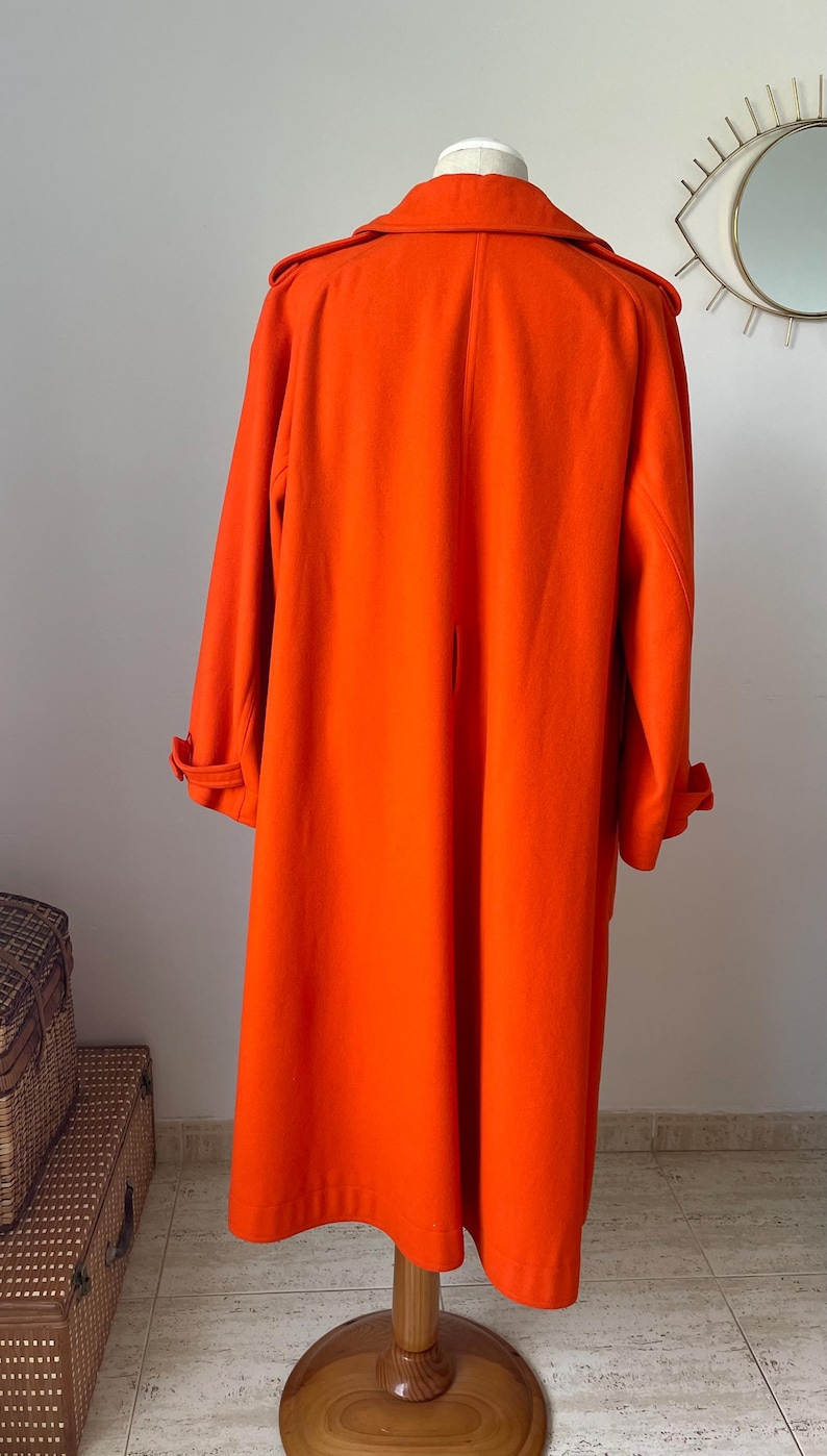 May include: Bright orange long coat with a classic design. The coat features a wide collar, long sleeves with buttoned cuffs, and a knee-length hem. It is displayed on a wooden stand, showcasing its full length and tailored fit.