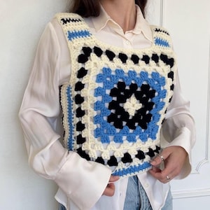 May include: A square-neck crochet vest in a granny square pattern. The vest features a central square of black, blue, and cream-colored squares. The edges and straps are trimmed with blue and cream crochet. The vest is worn over a white button-down shirt.