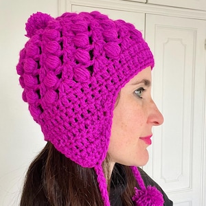 Puede incluir: Un gorro de ganchillo magenta vibrante con un patrón texturizado en forma de burbujas. El gorro presenta orejeras con borlas y pompones. Este accesorio de invierno hecho a mano es acogedor y elegante.