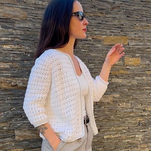 Puede incluir: Una chaqueta de ganchillo blanca con un patrón de punto calado texturizado, mangas tres cuartos y un dobladillo recto. La prenda presenta un ribete gris claro a lo largo de la abertura frontal y los puños, creando un contraste sutil.