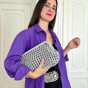 Puede incluir: Un bolso de mano tipo sobre tejido en crochet de color plateado metálico con un marco estructurado y un patrón calado. El bolso presenta un acabado brillante y reflectante con un cierre superior curvo y una textura artesanal detallada.