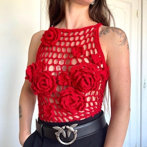 May include: A red crochet top with a net-like design, adorned with several three-dimensional rose appliqués. The top is sleeveless and has a high neckline. The roses are a vibrant red, contrasting with the open weave of the top.