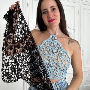 Puede incluir: Top halter azul claro con diseño floral de ganchillo. Un chal de ganchillo negro se sostiene frente al top. El top se combina con vaqueros azules. El tema general es la ropa hecha a mano.