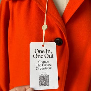 May include: Bright orange coat with a classic collar and black buttons. A white tag with the text "One In, One Out" and "Change The Future Of Fashion!" hangs from the coat. The coat is made of a soft, textured fabric.