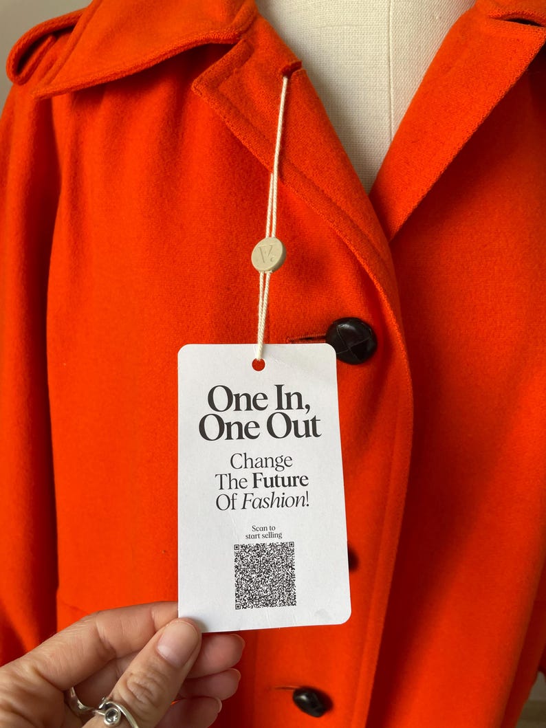 May include: Bright orange coat with a classic collar and black buttons. A white tag with the text "One In, One Out" and "Change The Future Of Fashion!" hangs from the coat. The coat is made of a soft, textured fabric.
