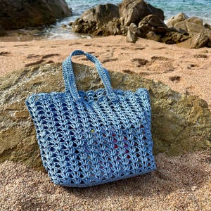 Puede incluir: Un bolso de mano de ganchillo en color azul claro con un patrón de punto calado, dos asas resistentes y un borde superior festoneado. El bolso está confeccionado con hilo texturizado y muestra un diseño geométrico repetitivo en el cuerpo.